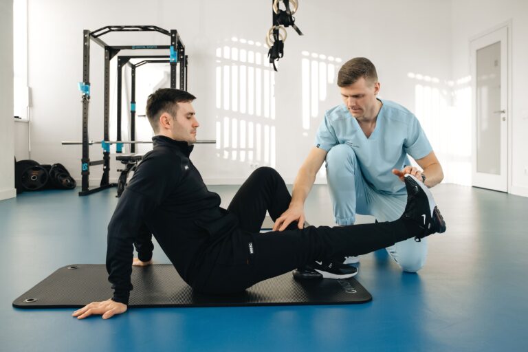 Fizjoterapeuta prowadzi ćwiczenia z pacjentem siedzącym na macie w sali rehabilitacyjnej